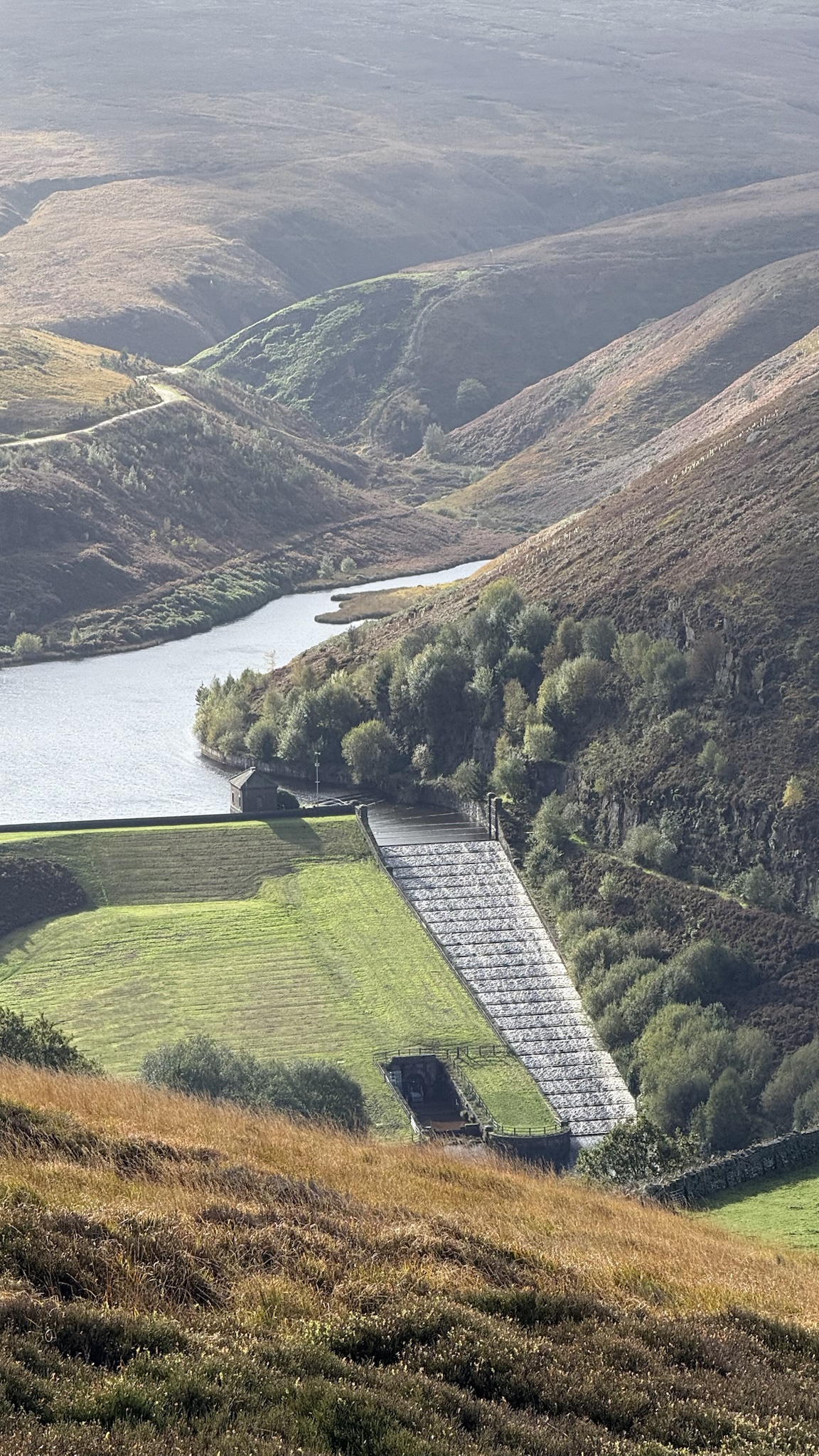 Looking down a steep moorland valley to a reservoir and a dam with a stepped stone spillway.